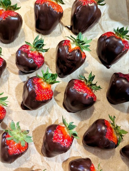 Hellbraunes, leicht verknittertes Backpapier, darauf viele, sehr große, dicke, rote Erdbeeren, die mit einer glänzenden, dicken, dunklen Schoko-Ganache überzogen sind. Nur rote Erdbeerenden und grüne Blättchen gucken heraus.