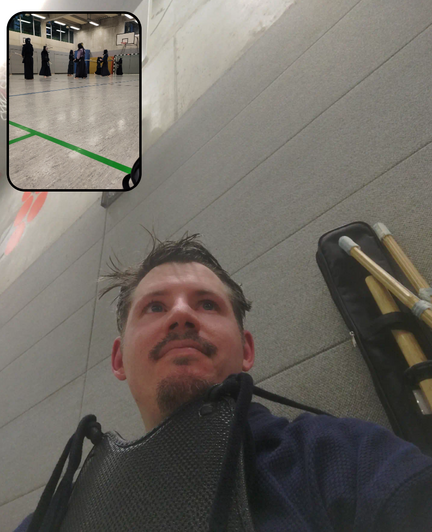 We see a man wearing is Kendō Armour except the Helmet called "Men". He is sitting in front of a grey wall of a sports gym hall. Behind him on the left you can see the tip of Bamboo swords called "Shinai". In the top left corner we see a picture in the picture depicting Kendō fighter in the mod of a practice. Fighting, clashing all while wearing Kendō armour.