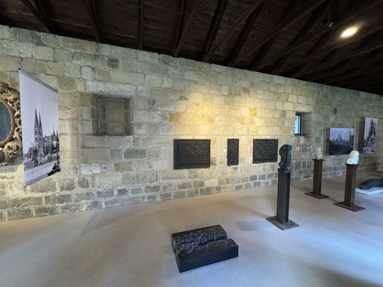 Blick in einen Raum, frontal ist eine Natursteinwand zu sehen, im Vordergrund ein heller Fußboden, in der Mitte sind schwarze und weiße Plastiken aus Stein in einer Ausstellung zu sehen, an den Wänden und quer im Raum hängen alte Bilder mit Motiven unmittelbar nach der Zerstörung Halberstadts 1945