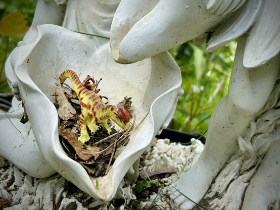 A green Dilophosaurus with brown spots stands in a dry flume clogged with dead leaves and twigs. A channel is above them with no running water. The fountain features are a white, faux porcelain.