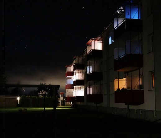 This image shows an apartment building at night. The building has several balconies, some of which are lit up with different colors of lighting. The sky is dark with visible stars, and there is some ambient lighting in the surrounding area.