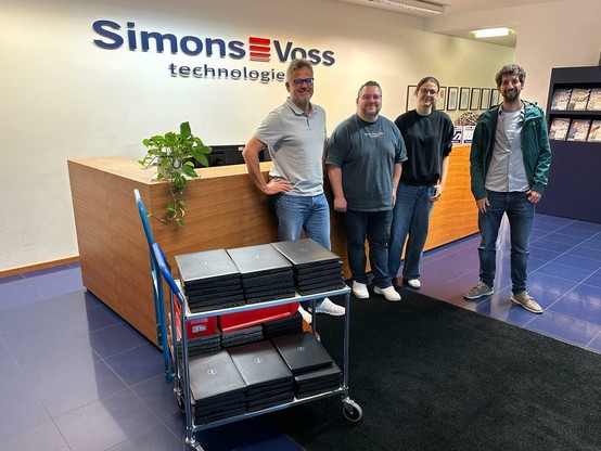 Am Empfang stehen 2 Mitarbeiter und 1Mitarbeiterin con SimonsVoss und Juan, Labdoo Hub München vor dem Tresen aus Holz. Auf einem Rollwagen vor ihnen stehen gestapelt 51 Laptops. Alle schauen lächelnd in die Kamera. Im Hintergrund das Logo von SimonsVoss auf einer weißen Wand.