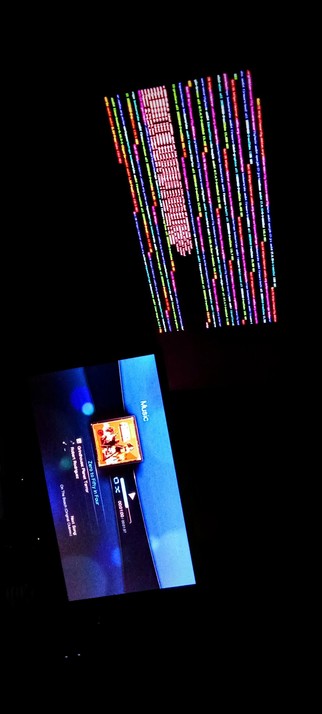 The photograph shot in total darkness is a split-screen view of two different displays. The top half shows a screen filled with colorful, vertical lines of text, resembling a computer terminal or a data stream. The colors include red, green, blue, and pink, and the text is arranged in a grid-like pattern, creating a vibrant and dynamic visual effect against a black background.

The bottom half displays a blue screen with a central image of a person, likely from a video game or media player interface. The screen shows the title "Zero to Fifty in Four" and the name "Robert Rodríguez," along with the text "On The Bench Original Soundtrack." The screen also includes a progress bar and a time indicator, suggesting it is a music player interface. The word "Music" is visible on the right side of the screen.

 Ovis2-8B

🌱 Energy used: 0.206 Wh