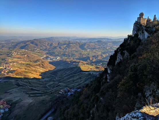 A scenic view of Guaita Tower atop Monte Titano in San Marino. The tower stands on a rocky cliff, overlooking a vast landscape of rolling hills and valleys under a clear blue sky. The foreground shows the rugged terrain leading up to the tower, while the background reveals the distant towns and the layers of hills fading into the horizon.