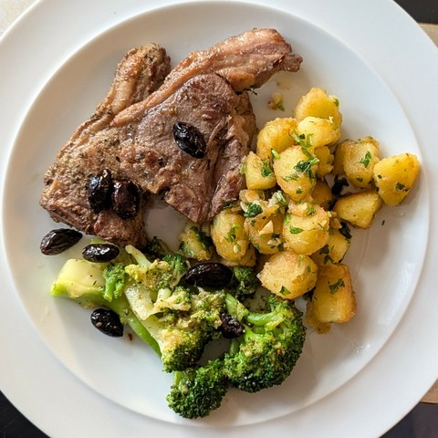 White plate, two small lamb chops, pile of small golden crisp potatoes dotted with garlic and parsley, broccoli, scattered black olives