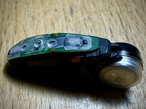 The top removed and now there’s another layer of “stuff”, this being clear flexible plastic that interfaces with rocker switch controls and maybe two screened openings where sound is guided to microphones below. There’s a thin green flexible circuit board below, but components are probably on the other side.