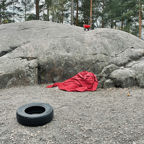 A playground scene in a forested area featuring a red blanket by a large rock, a tire lying on the ground beside it and a red little wheelbarrow above.