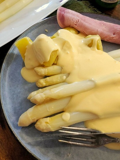 Esstisch, darauf ein großer, blaugrauer Teller mit Spargelstangen, Kartoffeln, einer Schinkenrolle und heller, cremiger Sauce über allem.