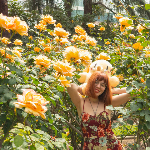 Photo of Lety and the Creature Mastodon stuffed toy standing in a garden bed surrounded by tall, yellow rose plants during a bright afternoon. Lety is shown wearing a red spaghetti strap dress decorated with floral designs. She's holding the Creature up on her shoulders behind her head, her palms behind its back, and her elbows in the air as she closes her eyes and smiles. The Creature is facing up toward the blooming Rosa “Autumn Gold” blossoms, the rose plants so tall that they stretch above the Creature and Lety's heads.