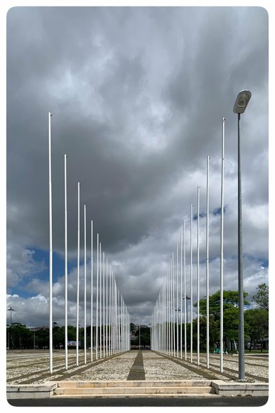 A long pathway lined with tall white flagpoles extends toward a cloudy sky, framed by greenery on either side. A streetlight and stone steps are visible on the right side of the image.