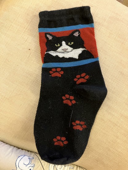 A black sock featuring a design of a tuxedo cat's face on the upper part, with red and blue stripes. The lower part of the sock has several red paw prints.
