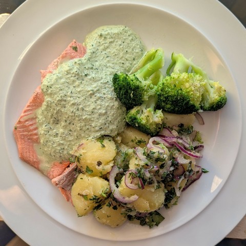 White plate, pinky/orange slab of salmon under pale creamy green sauce, pile of potatoes mixed with herbs and red onion, broccoli