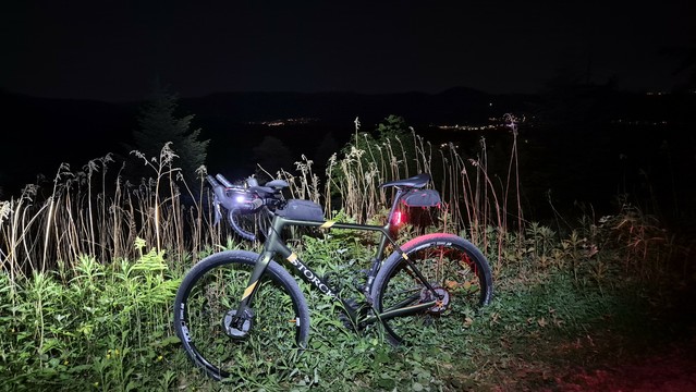 Ein grünes Gravelbike mit Beleuchtung steht nachts im Wald. Im Hintergrund erkennt man vereinzelt leuchtende Häuser in der Ferne in einem ansonsten schwarzen Tal.