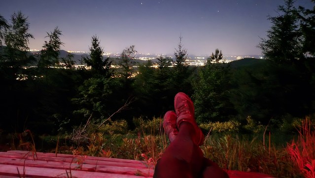 Eine Person liegt entspannt auf einer Holzbank im Wald und blickt über die nächtlichen Lichter im Tal.