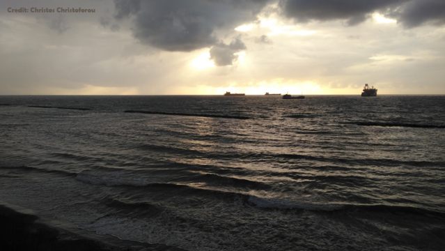 Limassol, Blick in die Akrotiri Bay. Der Himmel ist wolkenverhangen, dahinter leuchtet die Abendsonne durch, spiegelt sich auf leicht beweegter See. Vor der Küste liegen vier Handelsschiffe vor Anker. Ansonsten ist das Meer leer.
Links oben steht: Credit Christos Christoforou.