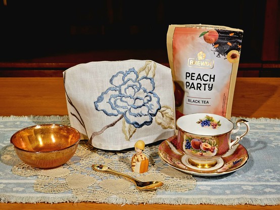 Tea tableau in tones of peach and sky blue, with vintage china with a a rich peach design, a carnival glass bowl, a miniature bust of a 1920s Flapper (from a collection of Avon bisque thimbles), a gold spoon with a cloisonne butterfly on the handle, an embroidered linen tea cozy, and Peach Party black tea from Brewix, all on a soft woven table runner graced by a fine beige lace vintage doily.