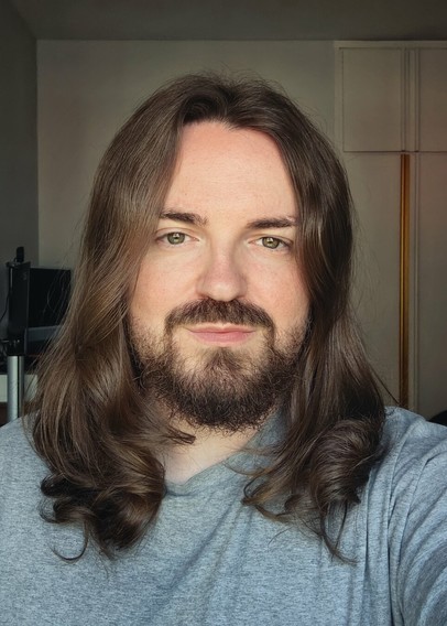 A selfie of me using a diffuser to reflect light into my face. I have long brown hair, light brown eyes, and a short brown beard.