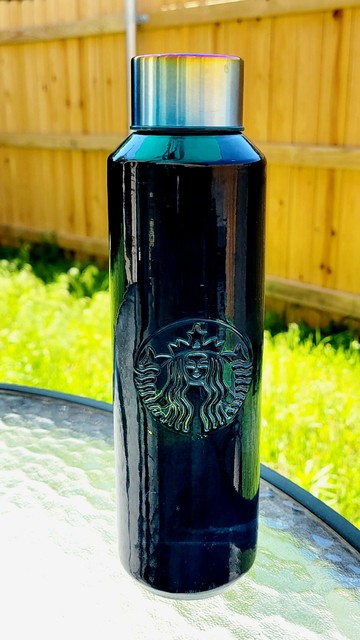 Blue-tinted glass water bottle with a chrome lid placed on glass tabletop in backyard. The bottle has the Starbucks siren logo.

Brown fencing in the background.