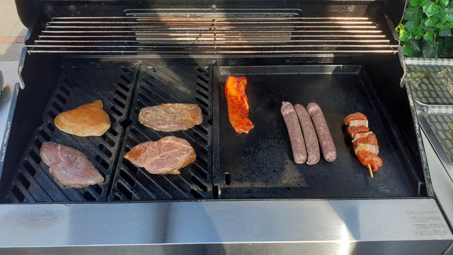 Ein offener BBQ Grill mit diversen Stücken an Fleisch und Wurst auf der Grillplatte und Rosten.