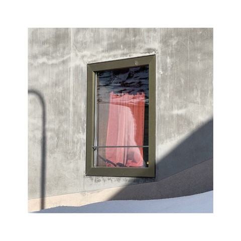 a single square window within a gray facade of a building. The curtain inside the window is red. alongside the left edge of the frame there's a curved shadow from a street lamp