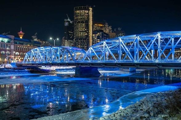 Photograph of iron girder bridge over a ice-locked river at night, with a city in the background.