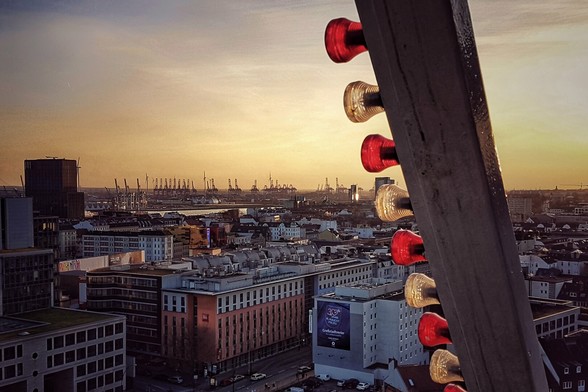 A high-angle view of a cityscape at sunset, taken from a Ferris wheel. The sky is a gradient of orange and yellow. City buildings and the dockland cranes are visible in the distance. The side of the Ferris wheel with illuminated red and clear lights is in the foreground on the right.