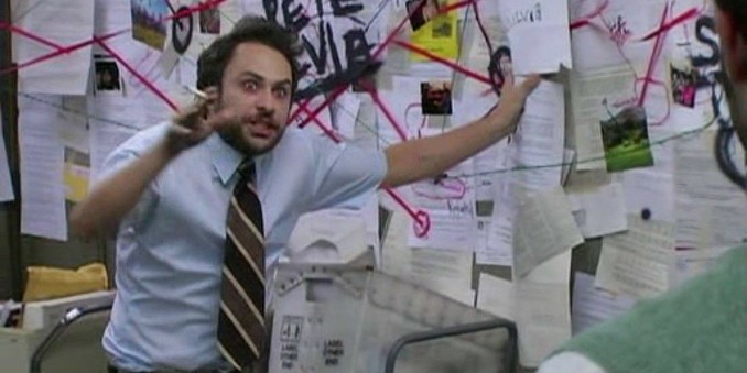A man with a beard, holding a cigarette, and excitedly pointing at papers tacked on a large wall board, with lots of red lines going from one piece of paper to another.