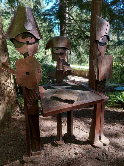 in a light & shade speckled forested area, a rusted iron sculpture stands, of three Picasso-esque masked musicians, holding a guitar, a recorder, and an accordion