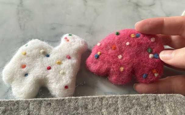 two needle-felted replicas of Mother’s Circus Animals cookies: a white camel and a pink bear, both with rainbow sprinkles. they’re about 2.5 times the size of actual cookies.