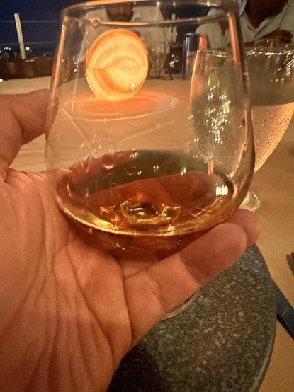A hand holding a glass of extra añejo tequila, with a blurred background featuring a soft glowing light and other glassware. The setting appears to be a dining area