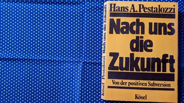 KI-generiert: Das Bild zeigt ein Buch auf einem dunkelblauen Stoff mit kleinen, weißen Sternen. Das Buch ist senkrecht aufgestellt und hat einen goldgelben Einband mit schwarzem Schriftzug. Der Titel lautet „Nach uns die Zukunft“ von Hans A. Pestalozzi.  Unter dem Titel steht in kleinerer Schrift „Von der positiven Subversion“.  Der Name des Verlages „Kösel“ ist sowohl am Buchrücken als auch unten auf dem Einband zu lesen. Der Stoff ist einfarbig dunkelblau und mit gleichmäßigen, kleinen weißen Sternen übersät, die ein gleichmäßiges Muster bilden. Das Buch liegt leicht schräg auf dem Stoff und nimmt etwa ein Drittel der Bildbreite ein.