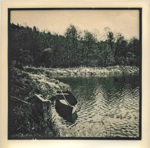 The image was printed using the lith print technique,  a photographic printing technique known for its distinctive high-contrast and grainy aesthetic, often with warm or sepia tones and dramatic shadow detail.

The image depicts a quiet, serene lakeside scene. In the foreground, a small rowboat is moored at the edge of the water, slightly tilted and partially reflected in the rippling surface. The shoreline is grassy and uneven, leading into a wooded area with tall, leafless or sparsely leafed trees, suggesting early spring. The dense forest in the background creates a dark, textured contrast to the lighter foreground. The overall mood is calm, nostalgic, and somewhat melancholic, enhanced by the lith printing process which gives the photograph a vintage, artistic look with rich blacks, soft highlights, and a warm tonal range.