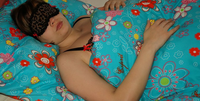 Das Bild zeigt eine Frau, die in einem Bett liegt. Sie trägt eine schwarze Schlafmaske mit einem leopardenartigen Muster und ist mit einem bunten Bettlaken bedeckt, das mit großen, farbenfrohen Blumenmustern verziert ist. Das Bettlaken ist in einem leuchtenden Blau gehalten und zeigt verschiedene Blumen in Rot, Gelb, Grün und Weiß. Die Frau liegt auf dem Rücken, mit einem Arm unter dem Kopf und der anderen Hand auf dem Bettlaken. Sie trägt ein schwarzes Spitzen-BH. Die Gesichtszüge der Frau sind teilweise verdeckt durch die Schlafmaske, aber sie scheint zu schlafen. Die Umgebung wirkt gemütlich und entspannt.
