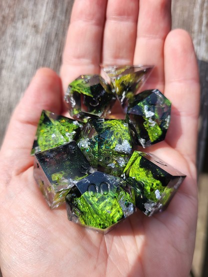 The palm of a hand holding a TTRPG dice set with bright green snake skin, silver foil and glittery dark green swirls.