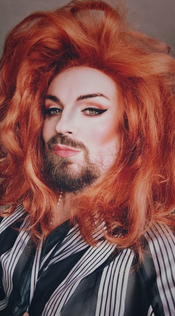 A highly stylized selfie with exaggerated contrast and saturation. I am wearing a fairly messy ginger wig that is shoulder length, and a silver and black striped shawl. My face is covered in makeup with very defined eye and cheeks, and pink corally lipm.