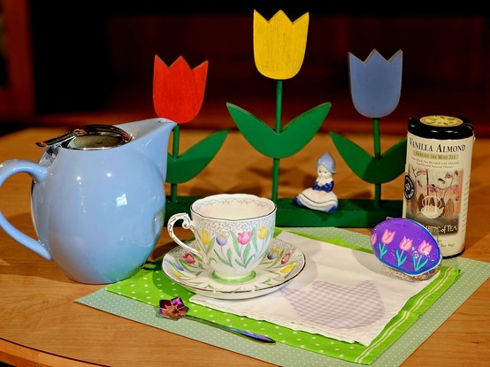 Tea tableau on a green ground with a white cloth appliqued with a lavender gingham tulip. Upon this are a vintage cup and saucer set patterned with a row of pastel tulips. Beside it are a handpainted purple rock with pink tulips, a tin of Vanilla Almond black tea, a powder blue teapot, and an iridescent purple spoon shaped like a flower. Behind them is a wooden sculpture of bright tulips in red, yellow, and blue, and a small Delft figurine of a little Dutch girl rests on the base.