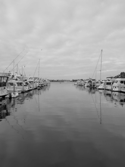 Black and White picture of the San Diego Marina on a cloudy day. On Each side of the frame, boats are lined up and in the center of the channel, with the North Island in the background, a small with 2 people fishing