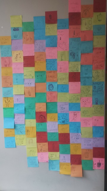 Multi-coloured mosaic of Post-It notes, each with doodles by children, covering the wall of the local bubble tea cafe