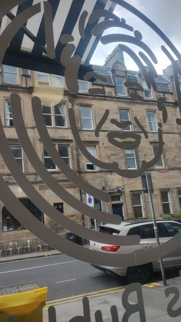 Looking through the window of Jan's Bubbles, with the outline of a smiling face drawn on the glass, a Victorian stone tenement visible opposite