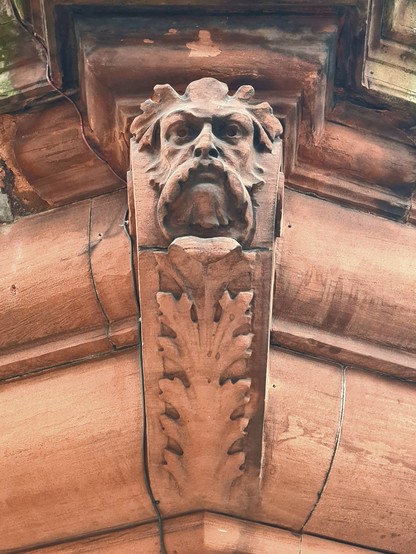 A sculpted corbel on a red sandstone Victorian building with a mask of a green man at the top.