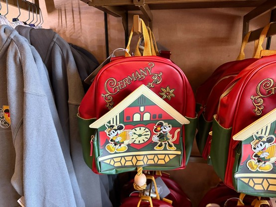 Red and green backpack with "Germany" text and cartoon mice by a cuckoo clock, hanging on a store display.