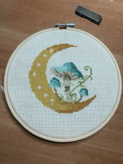 A round cross stitch piece depicting blue mushrooms on a white field with a yellow crescent moon.
