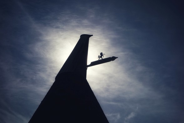A low-angle, silhouetted view of a traditional oast house against a bright, cloudy sky. The conical roof of the oast house dominates the left side of the frame, leading up to a cowl. A weather vane, featuring a small horse figure, extends horizontally from the side of the cowl towards the right. The sun is positioned behind the oast house and clouds, creating a bright halo effect and silhouetting the structure and the weather vane. The sky transitions from a bright, almost white area behind the oast house to a darker, more textured blue in the upper and right portions of the image.
