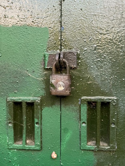 A rusty padlock is secured on a green metal surface, with two slots on either side of the lock. A small snail is resting near the bottom of the image. The surface appears weathered and slightly textured.