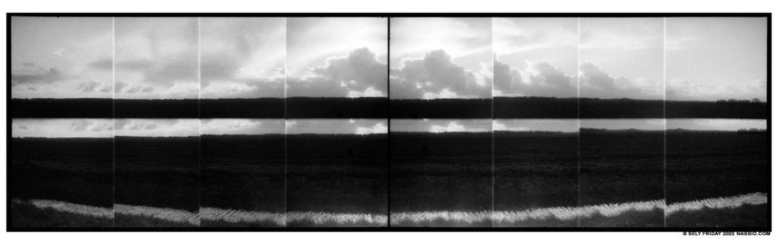 Black and white panorama photograph comprised of 16 individual shots in camera of the sun just behind some puffy clouds off in the distance, above a polder. Just in front of the polder is a reflecting body of water.