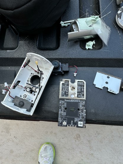 An assortment of electronic components, including a disassembled device casing, a circuit board, and some loose parts, are scattered on a surface. A shoe partially visible at the bottom adds context.