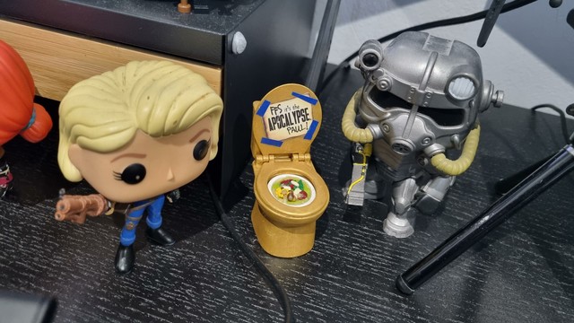 Two fallout themed funkopops - left is a vault dweller with blonde hair, right is a power armor - either side of a golden toilet with soup in the bowl and the words "FFS it's the apocalypse Paul!" Written on the lifted lid