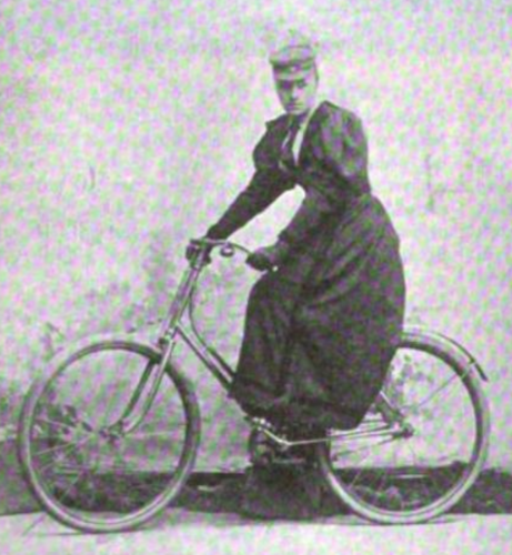 Ida Branth, on a bicycle, from an 1890s publication; she is wearing a small peaked cap and a dark dress with puffy sleeves and a long skirt; also dark gloves