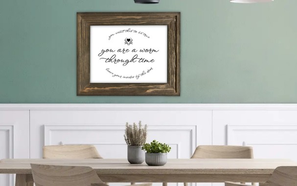 Dining room stock photo featuring a light, natural wood table with two small houseplants on it and matching chairs. The wall behind the table is white with decorative moulding, then halfway up, the wall becomes flat and green. A brown wooden picture frame on the wall contains the focus of the image, fancy black cursive lettering on a white background. The center, in large print, reads "you are a worm through time". Above it is a heart with lines splaying outward from it and a line of text, curved upward, which reads "you want this to be true". Below, a line of text curved downward reads "leave your insides by the door".