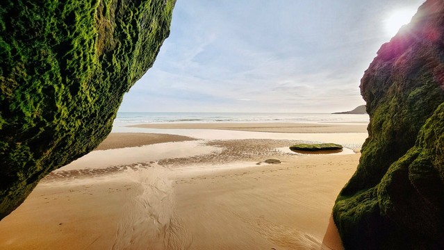 A scenic view of a sandy beach and calm ocean, framed by two large, moss-covered rocks in the foreground. The sky is partly cloudy with the sun shining brightly.